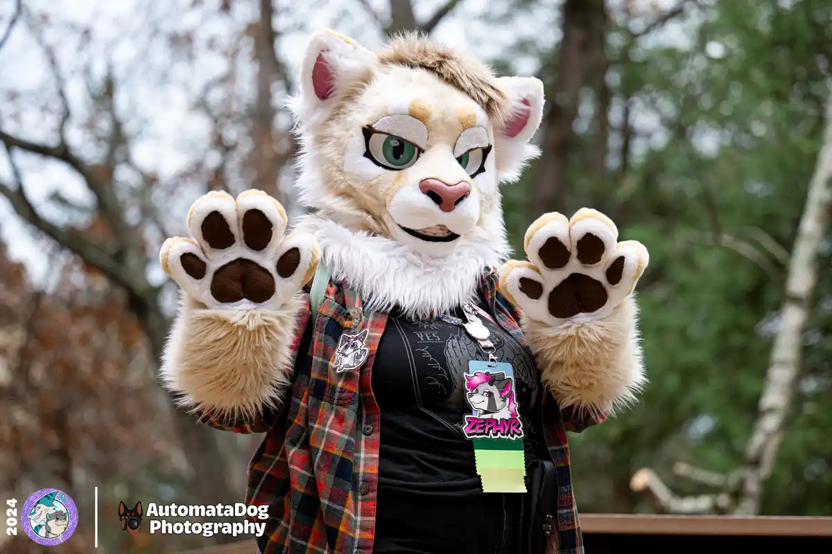 A person in a detailed fursuit of a tan and white cat-like creature with green eyes and paw gloves holds both paws up in a…