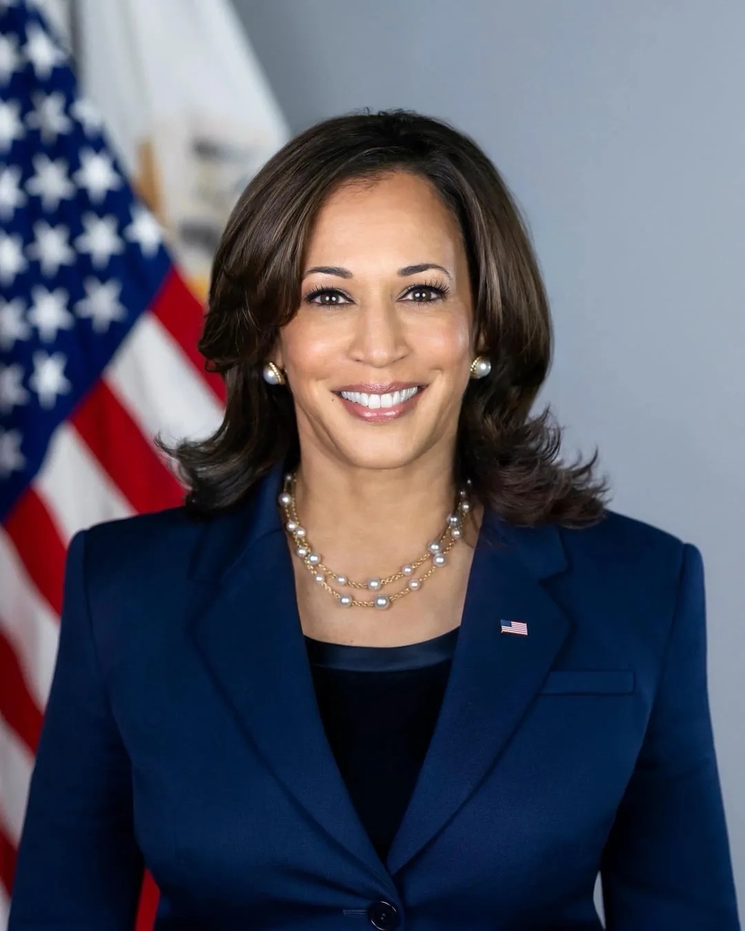 a formal portrait of Kamala Harris, smiling, wearing a dark blue suit with a pearl necklace and American flag pin, standing…