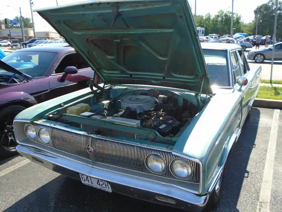 a light blue vintage car with its hood open, revealing an old engine and visible rust, parked in a lot with other cars. The…