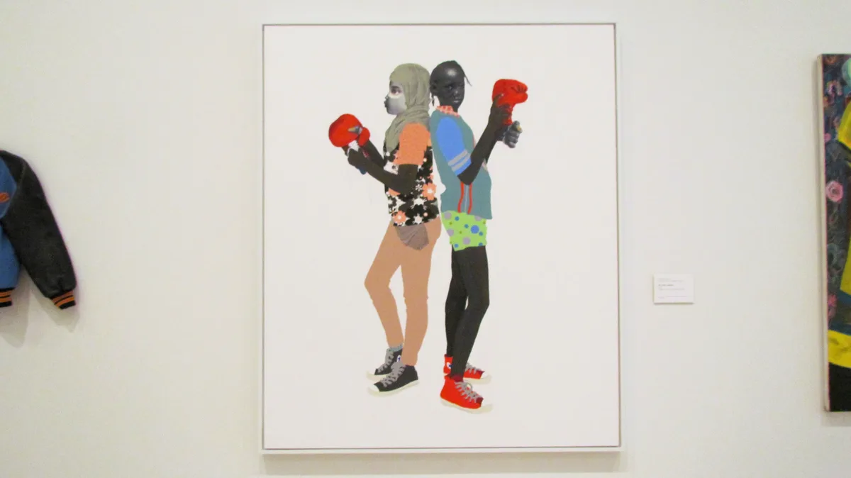 A framed painting in a gallery shows two young people standing back-to-back, both wearing red boxing gloves. The person on…