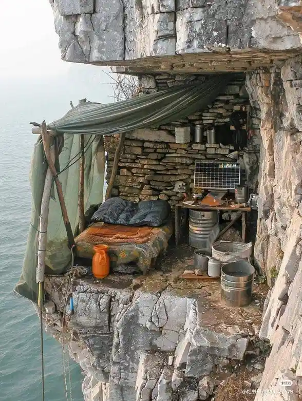 A rustic, makeshift shelter built into a cliffside overlooking water, featuring a bed with blankets, a green tarp roof, and…