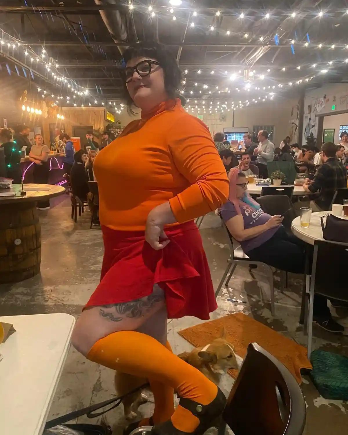 A person in a bright orange turtleneck, red skirt, and orange thigh-highs poses confidently in a restaurant with string…