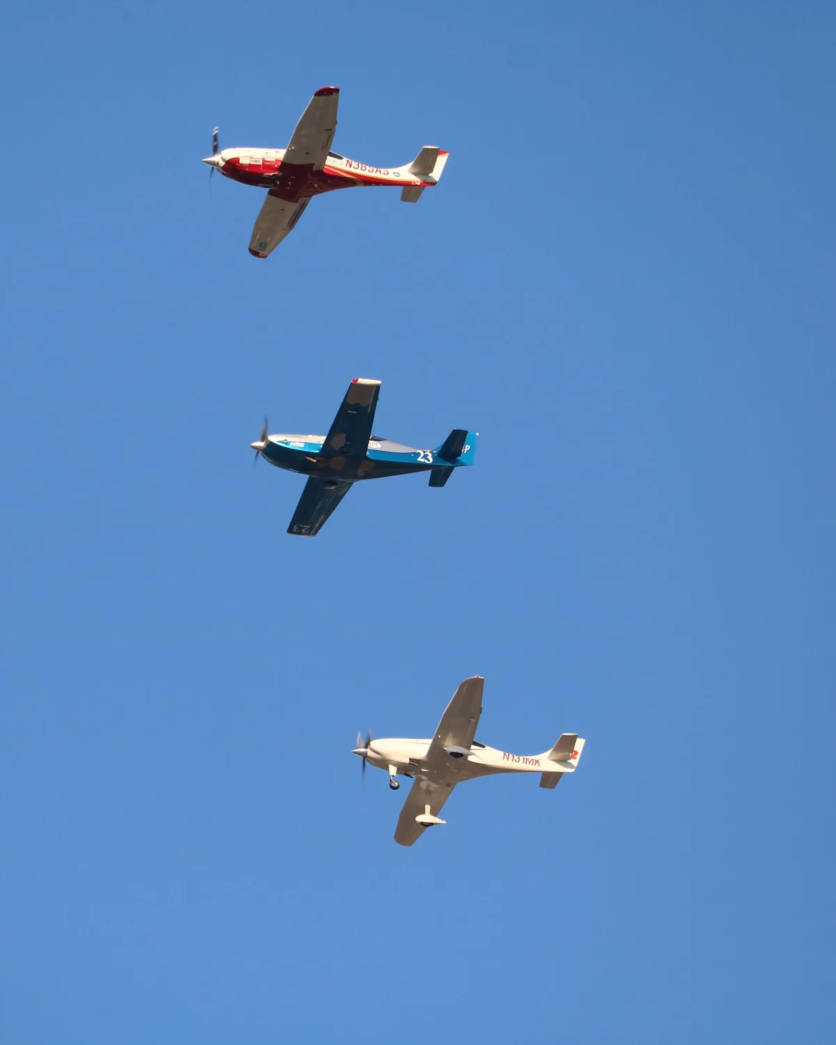 Three small propeller planes fly in a vertical line against a clear blue sky. The top plane is red and white, the middle one…