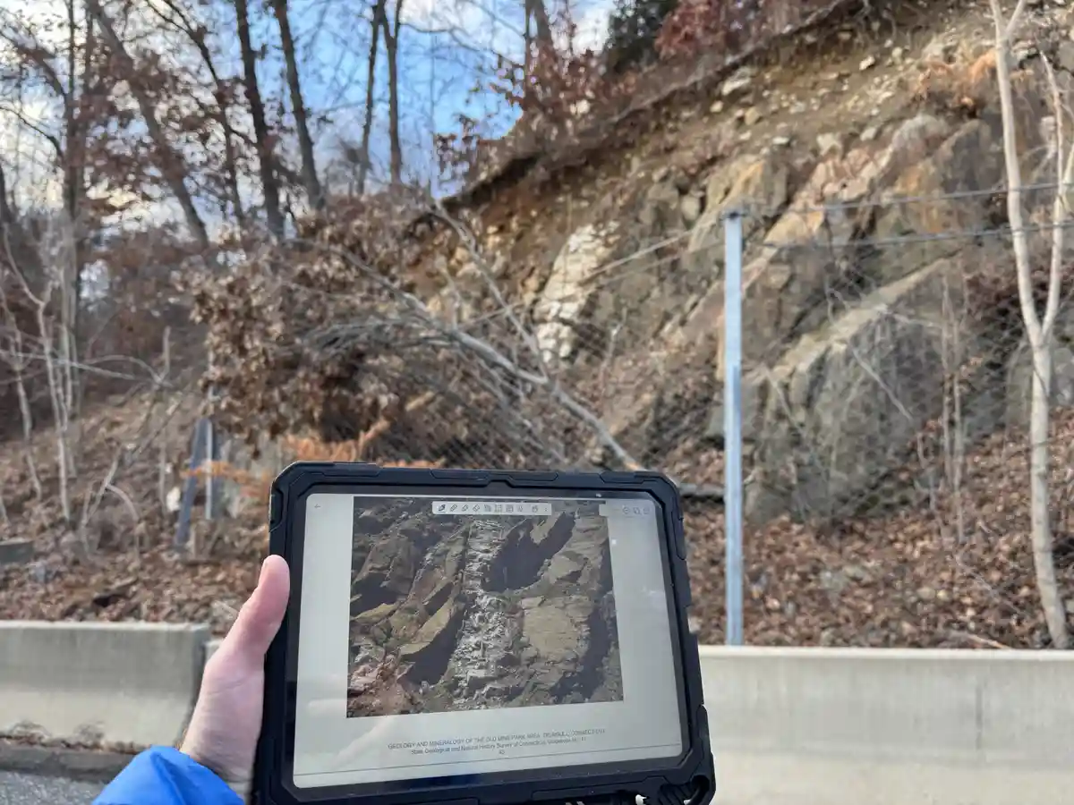 A person holds a rugged tablet displaying a geological guidebook image of a rocky cliff face, standing in front of the…