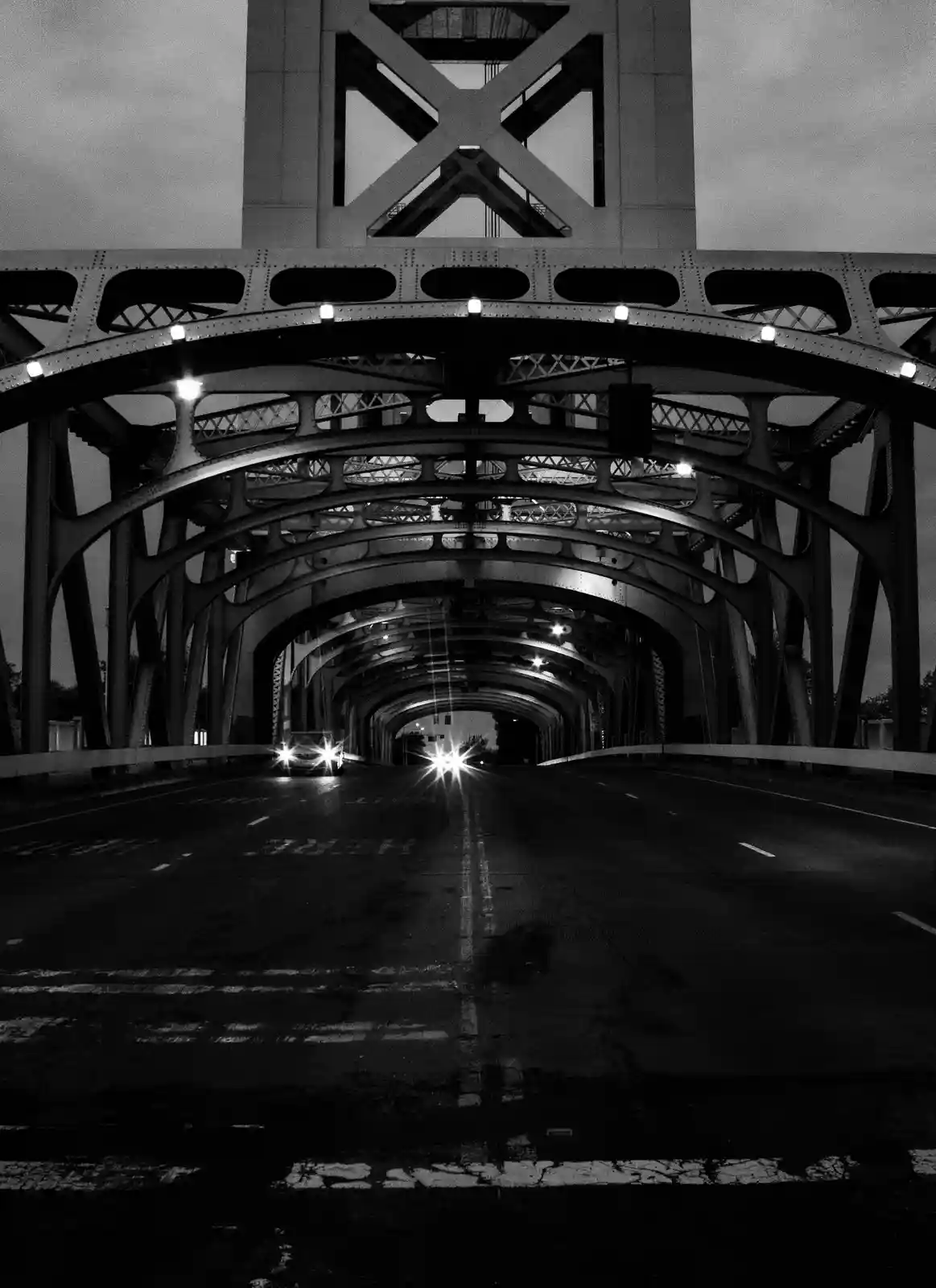 A black-and-white photo shows a low-angle view from inside a large, arched steel bridge at night, with two cars approaching…