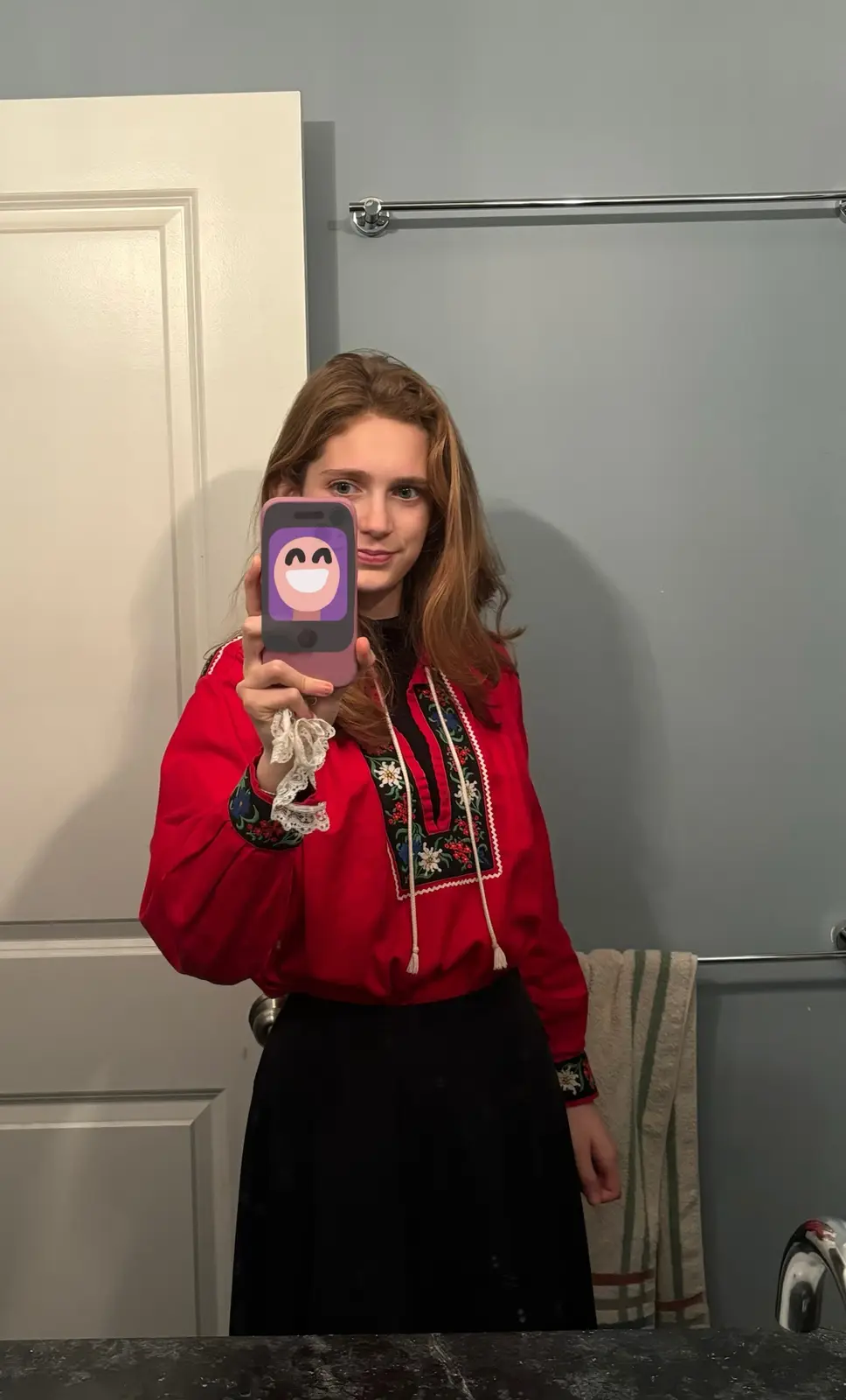 A young woman with brown hair stands in a bathroom mirror selfie, wearing a red embroidered blouse and black skirt. She…