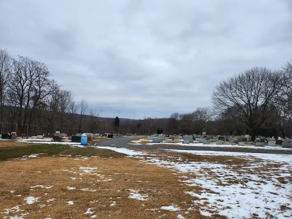 A wide shot of a cemetery in winter with patches of snow on brown grass, numerous headstones, bare trees, and wind turbines…