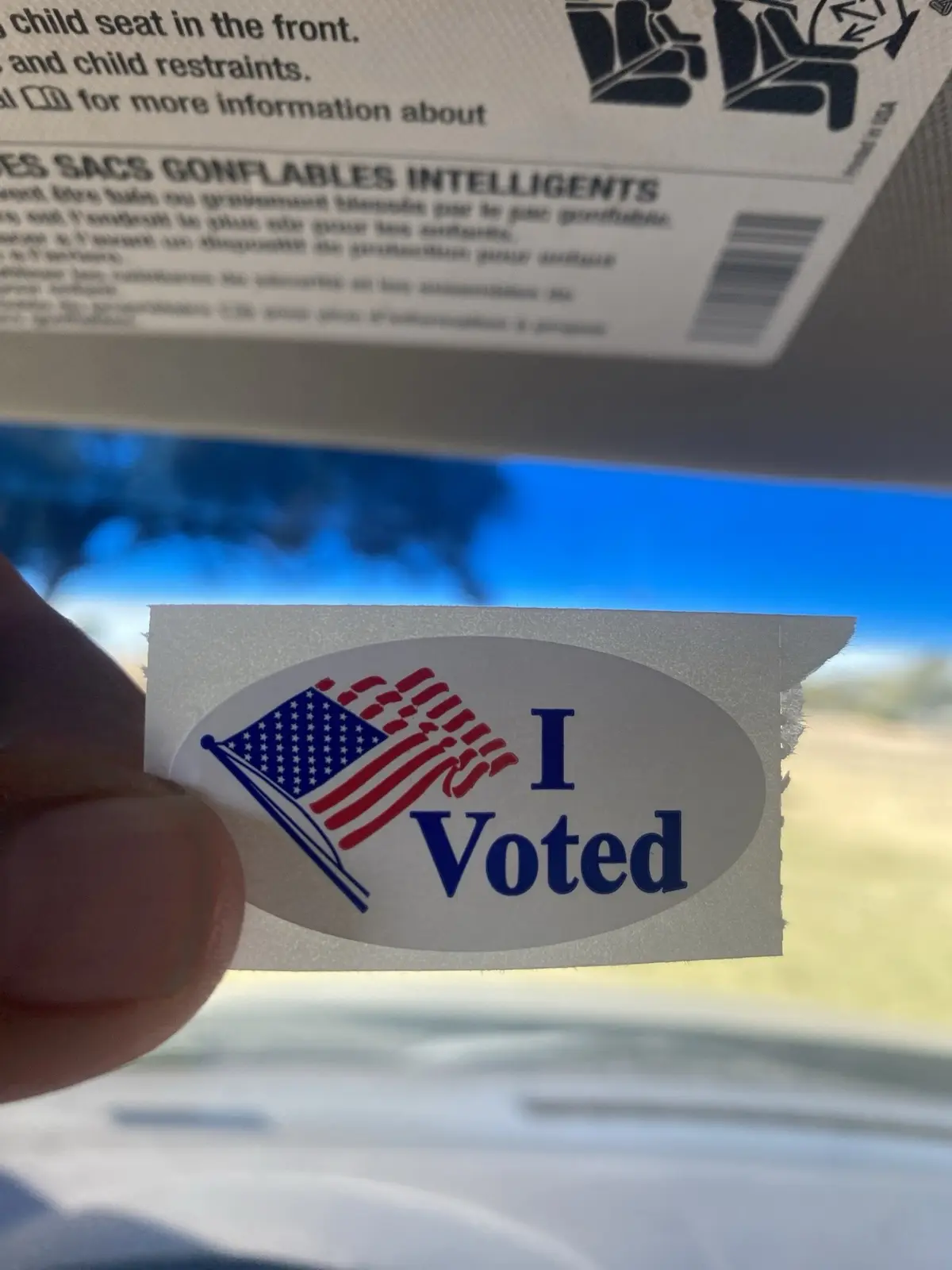 A hand holds up a torn 'I Voted' sticker featuring an American flag, inside a car with a visible sunroof and safety…