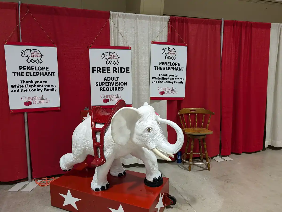 A white plastic elephant ride, named Penelope, stands on a red platform with white stars. Three signs hang behind it: two…