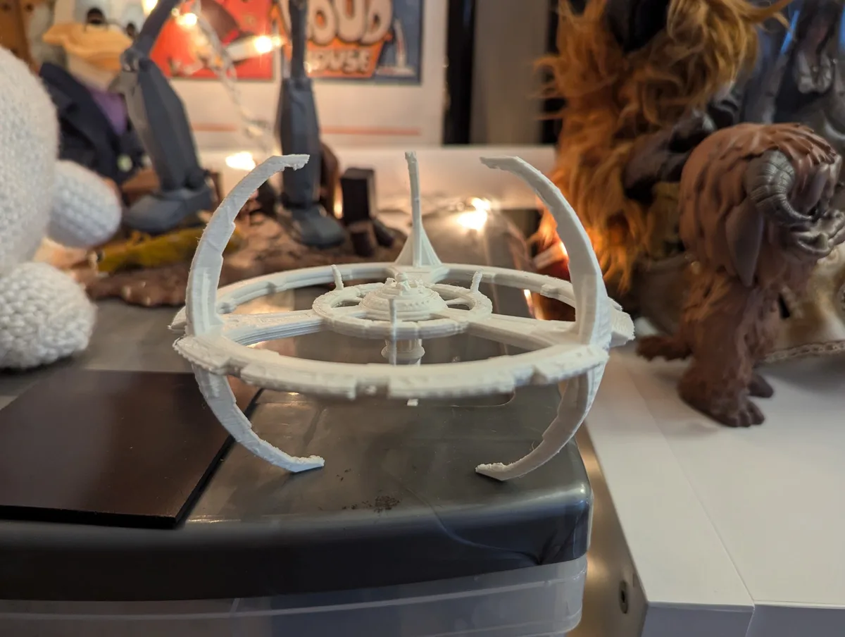 A white 3D-printed model of a Star Destroyer sits on a plastic storage bin, surrounded by other figurines including a…