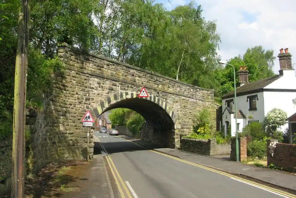 A stone archway over a road in a rural area, with a triangular '12'3"' height restriction sign mounted on it. A 'School'…