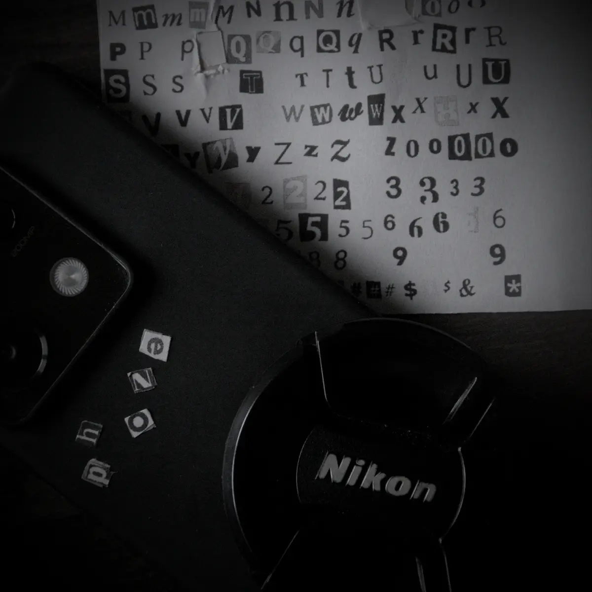 A black smartphone with a visible camera module and '200MP' text lies next to a Nikon lens cap, with scattered letter tiles…