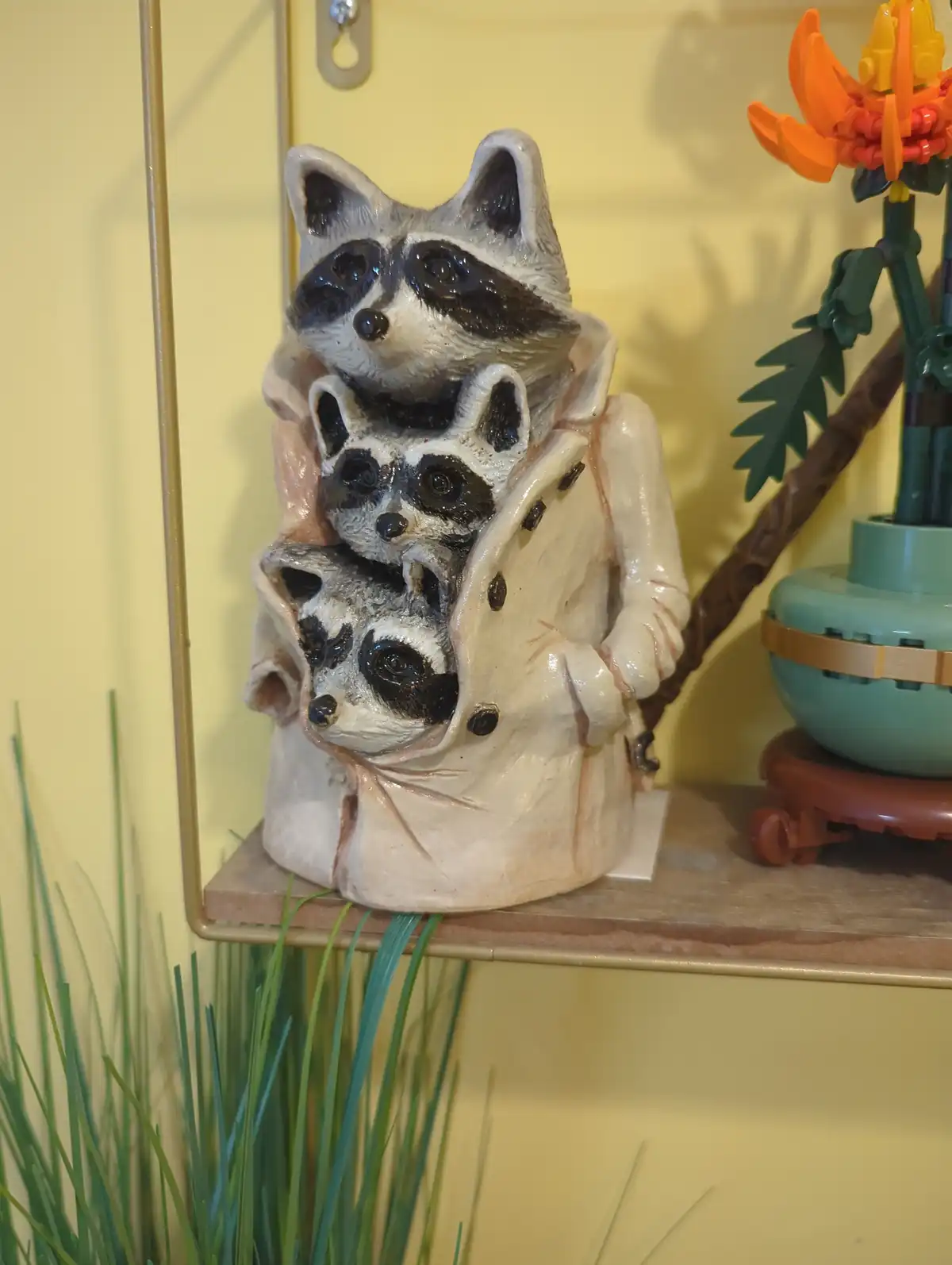 A ceramic figurine shows a raccoon wearing a trench coat, with two smaller raccoons peeking out from inside the coat. The…