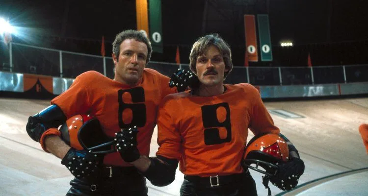 Two men in orange football jerseys with the numbers 6 and 9 stand together on a rink, one with his arm around the other. The…