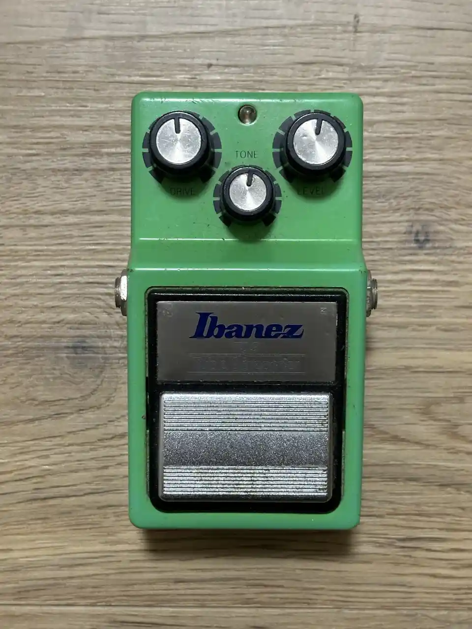 A green Ibanez TS9 Tube Screamer guitar effects pedal with three knobs labeled DRIVE, TONE, and LEVEL sits on a wooden…