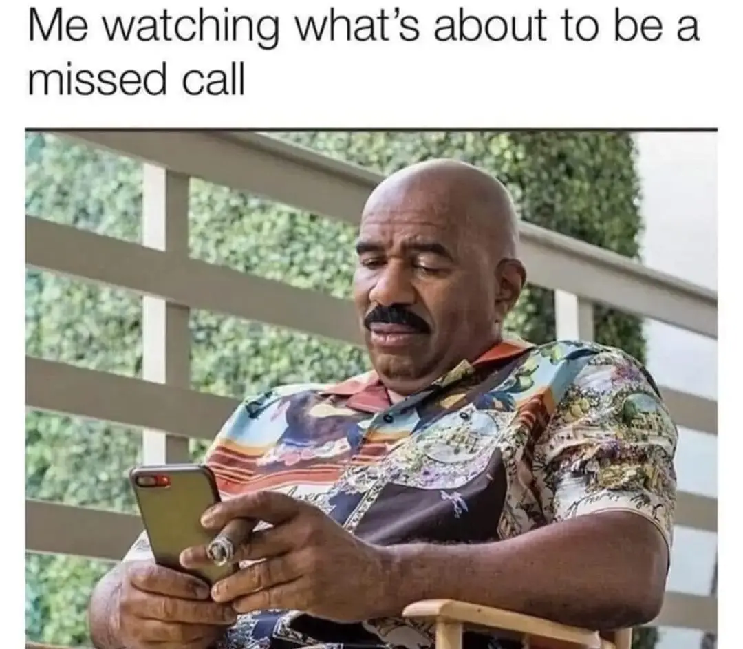 A bald man with a mustache wearing a colorful Hawaiian shirt sits in a chair holding a smartphone in one hand and a cigar in…