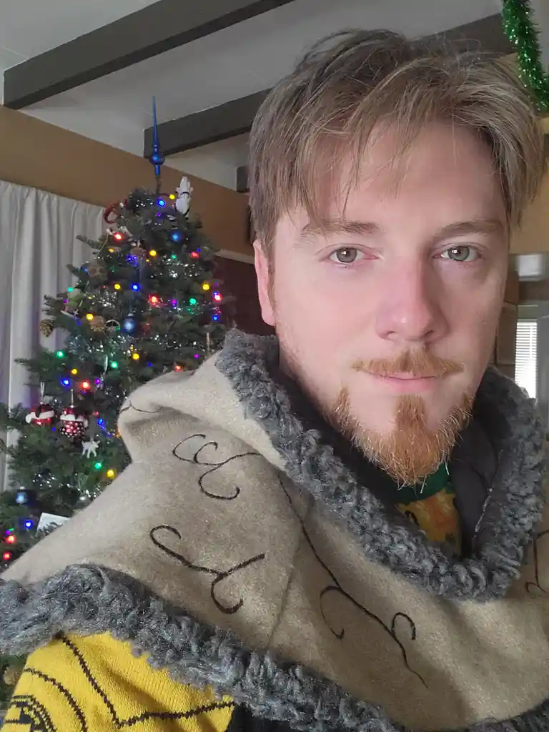 A man with reddish-blond hair and a goatee looks directly at the camera with a tired, slightly smug expression while wrapped…