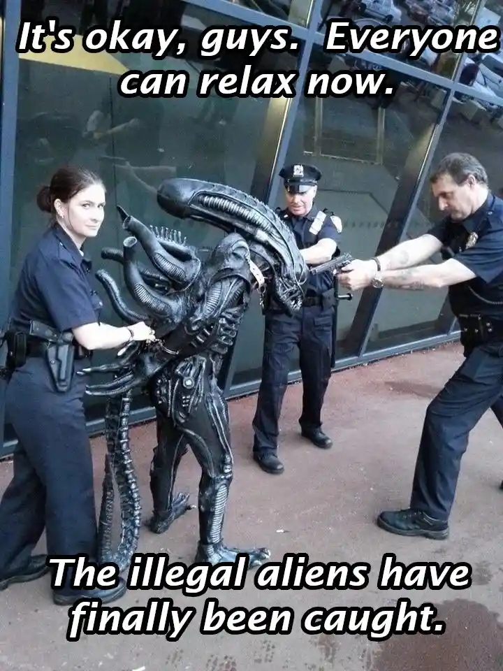 Three police officers are surrounding a person in a full-body Alien costume, with one officer holding a gun pointed at the…