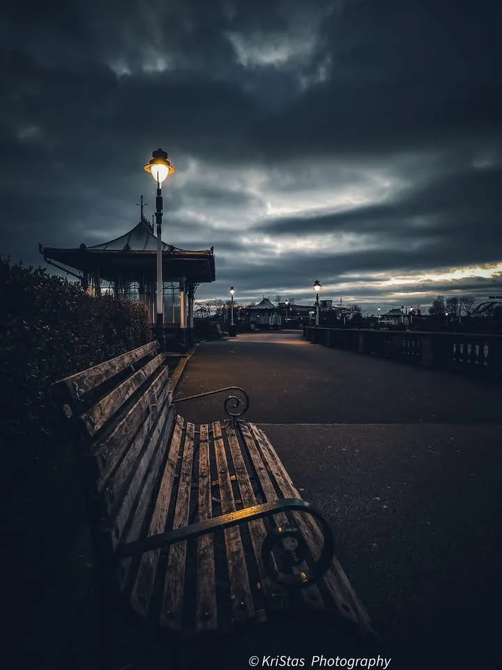 A dimly lit, atmospheric photograph of an empty wooden park bench under a gloomy, cloudy sky, with a single illuminated…