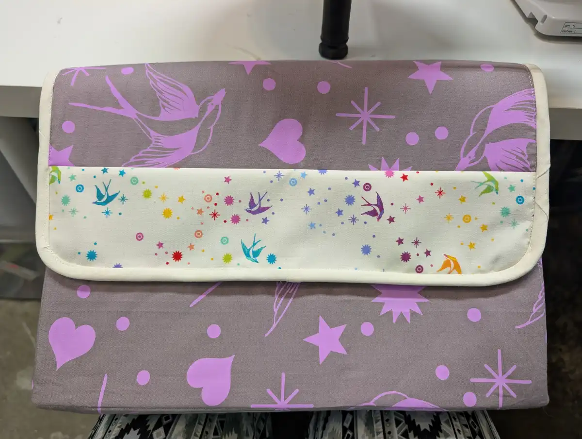 A handmade fabric clutch or pouch with a purple and grey pattern featuring birds, hearts, and stars. The flap has a…