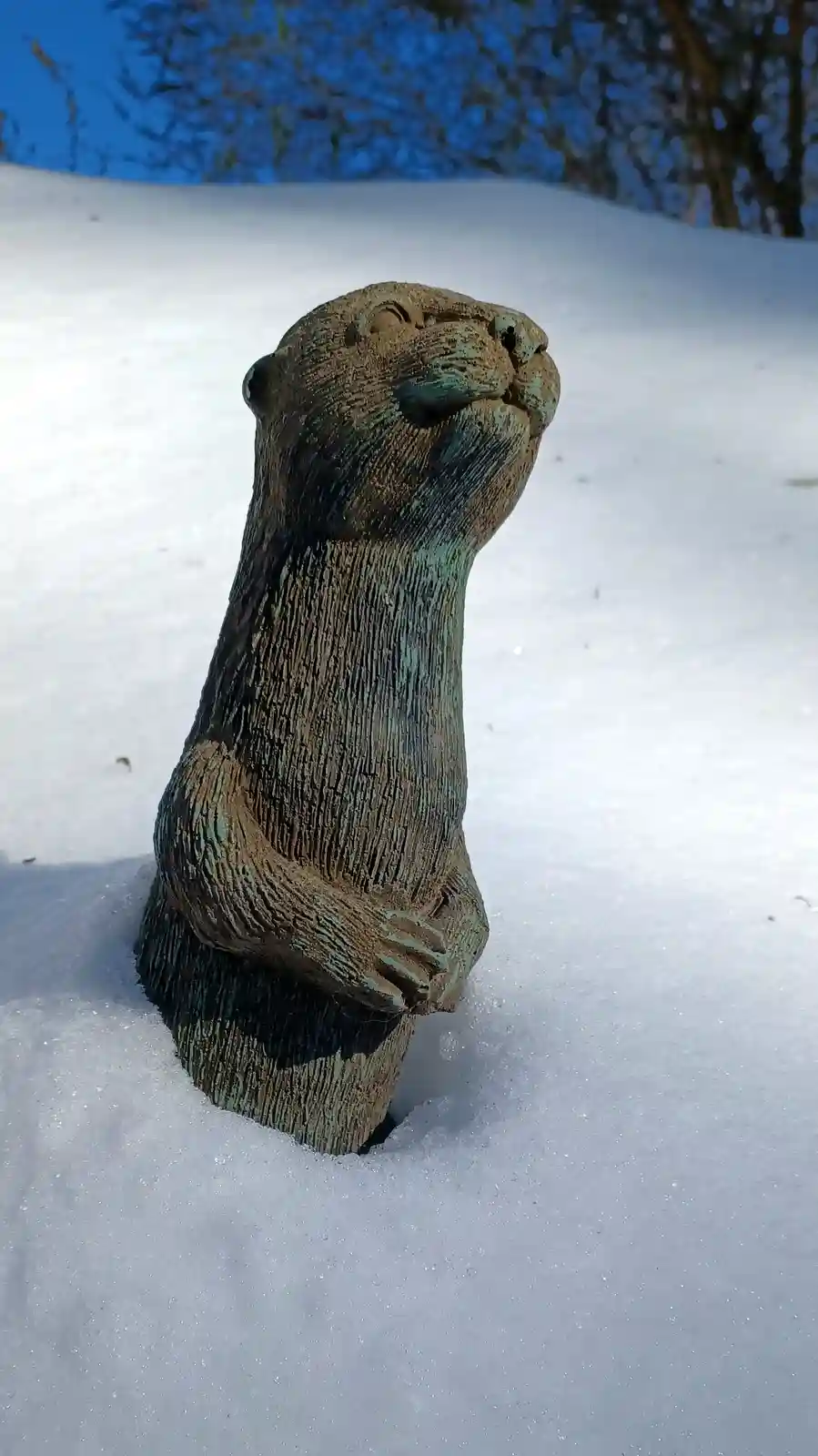 A weathered wooden otter statue, partially buried in snow, looks upward with a serene expression. The overlaid text reads…
