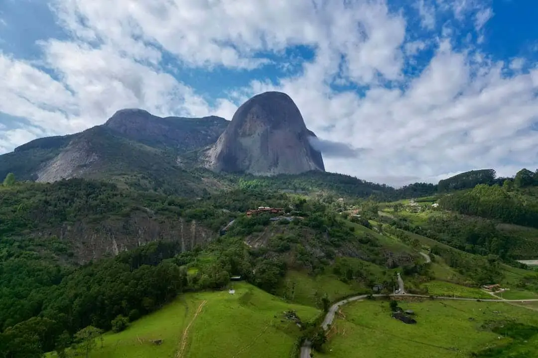 A scenic landscape photo of a large, rounded mountain dominating a green, forested valley under a partly cloudy blue sky. A…
