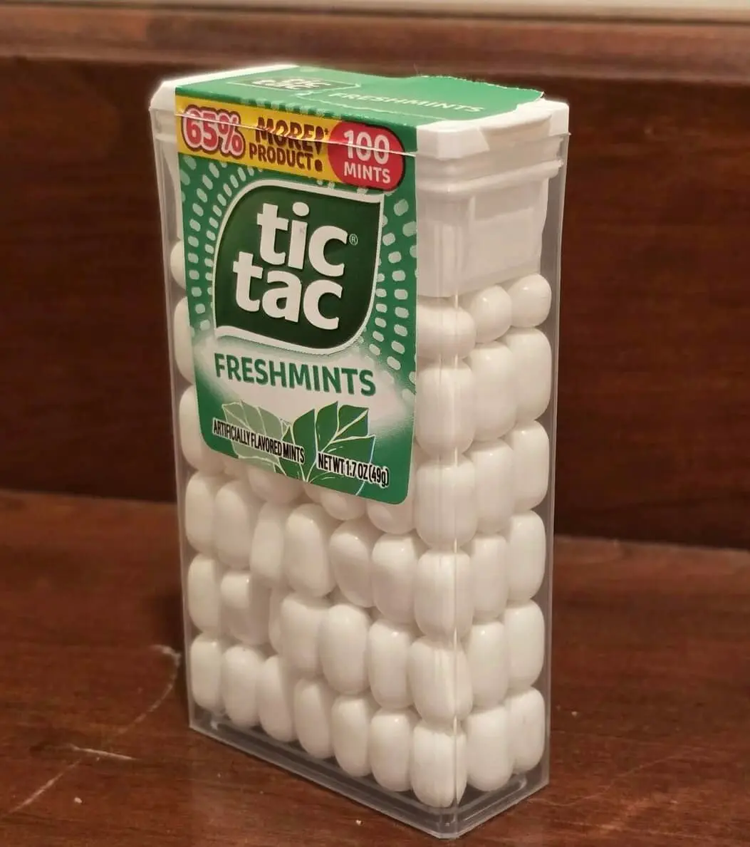 A clear plastic container of Tic Tac Freshmints sits on a wooden surface, filled with white mints. The green and white label…