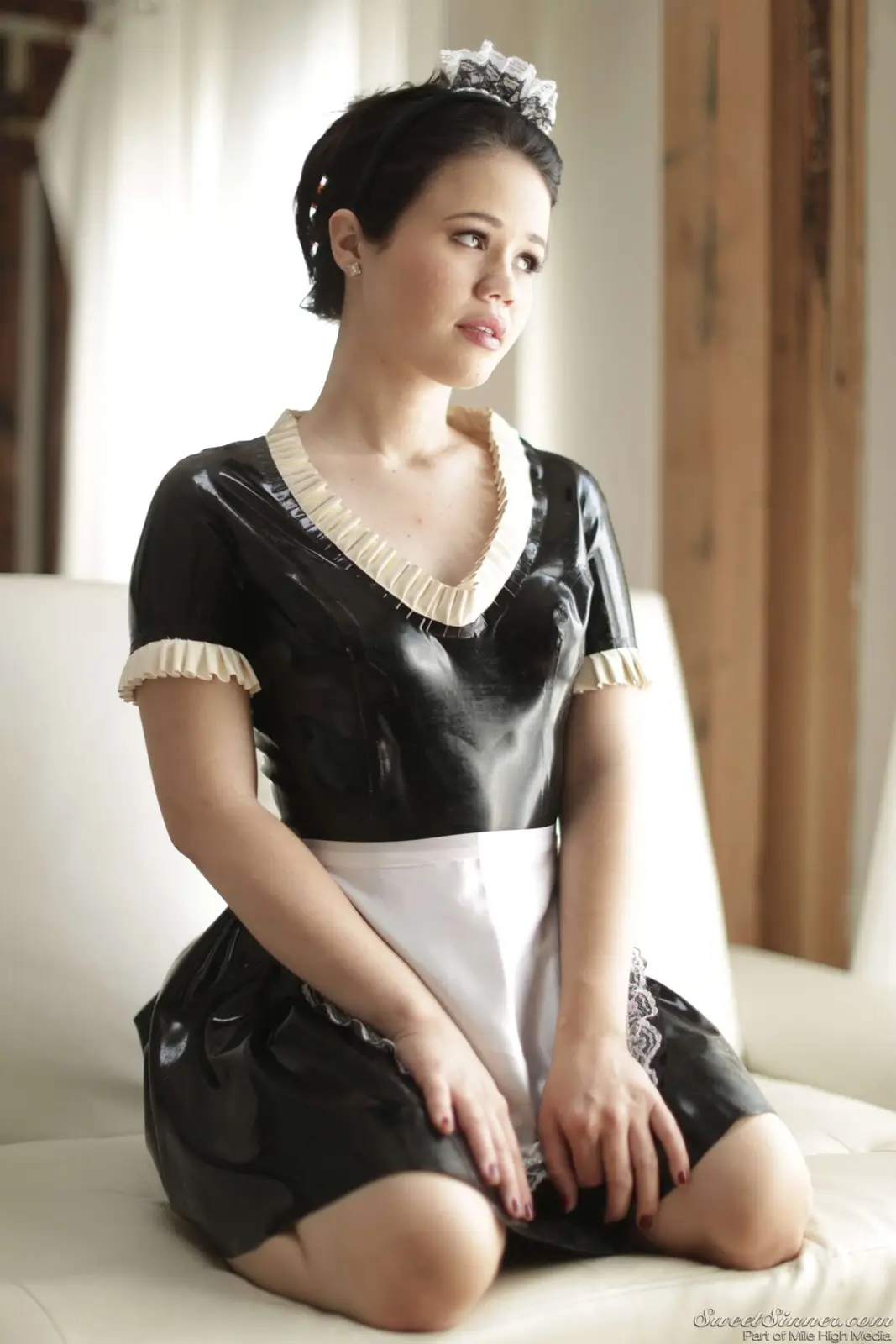 A woman in a black latex maid outfit with white frills and a lace headband sits on a white couch, looking off to the side…