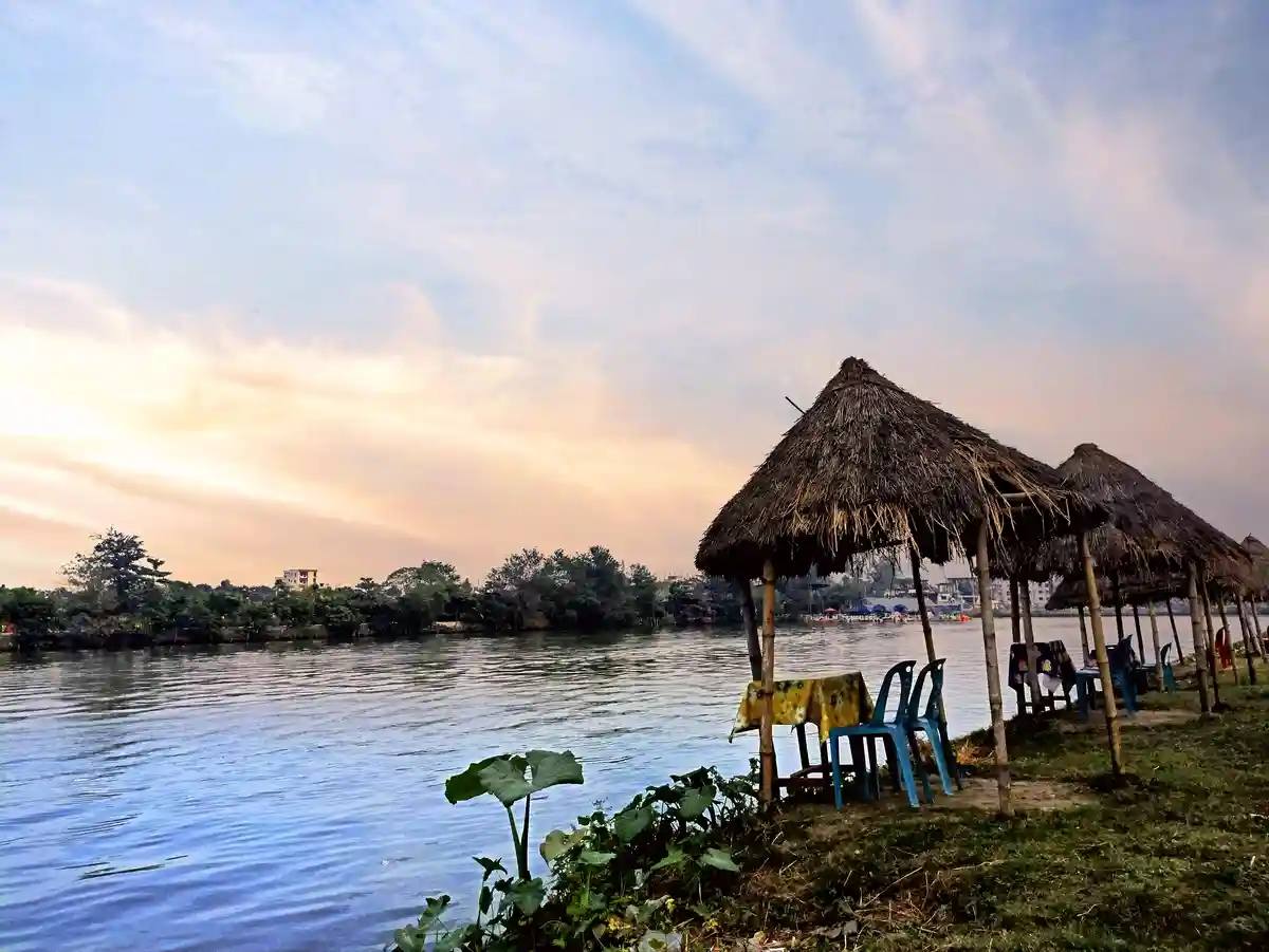 A serene riverside scene at dusk with thatched-roof huts, blue plastic chairs, and tables on a grassy bank. The sky is…
