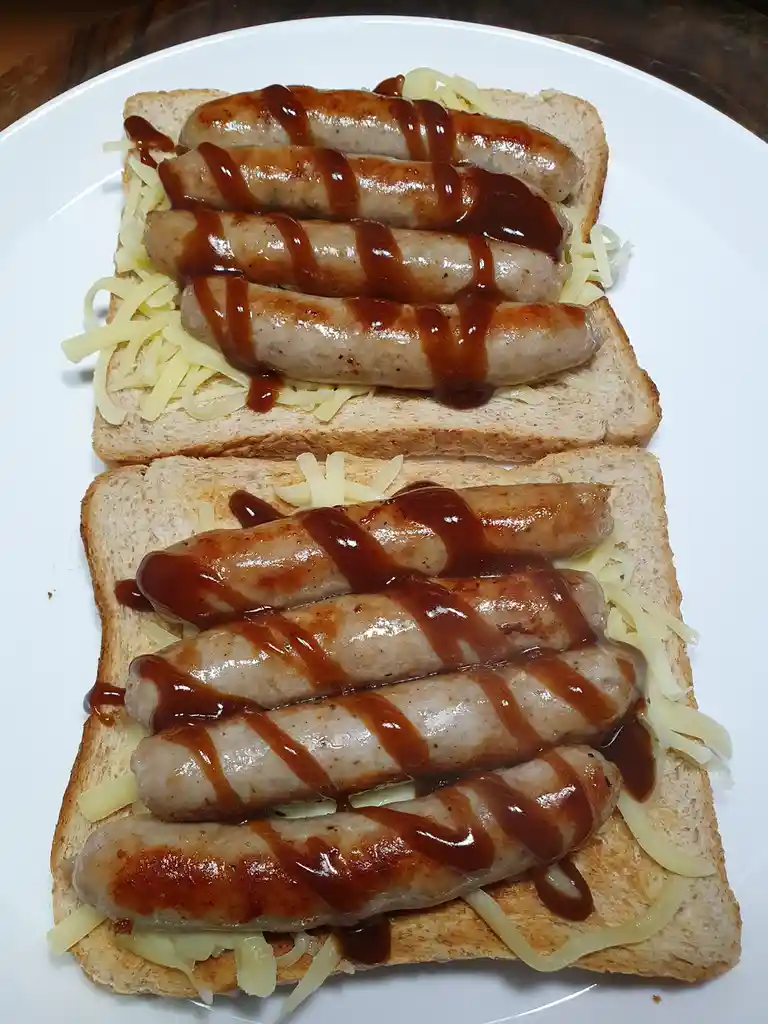 Two slices of toasted bread are topped with shredded cheese and four grilled sausages each, drizzled with a dark sauce…