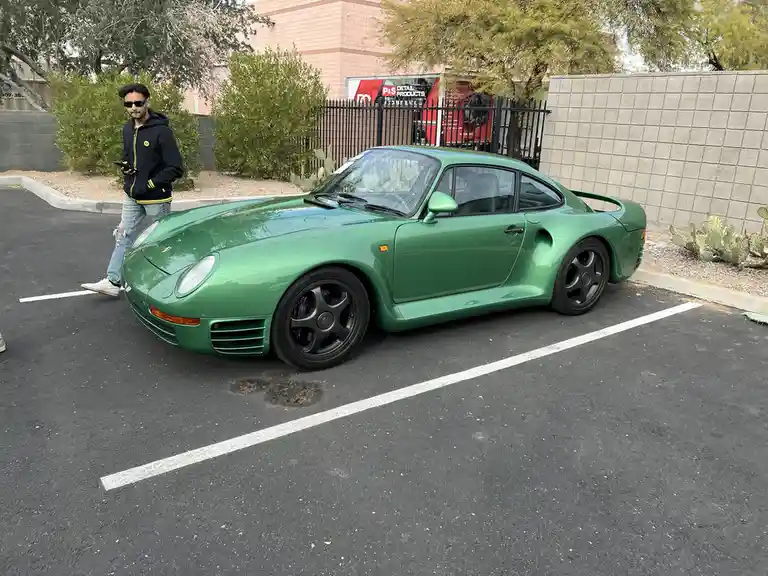 A man in a black hoodie and sunglasses walks past a shiny green Porsche 959 parked in a lot, looking at his phone. The car…