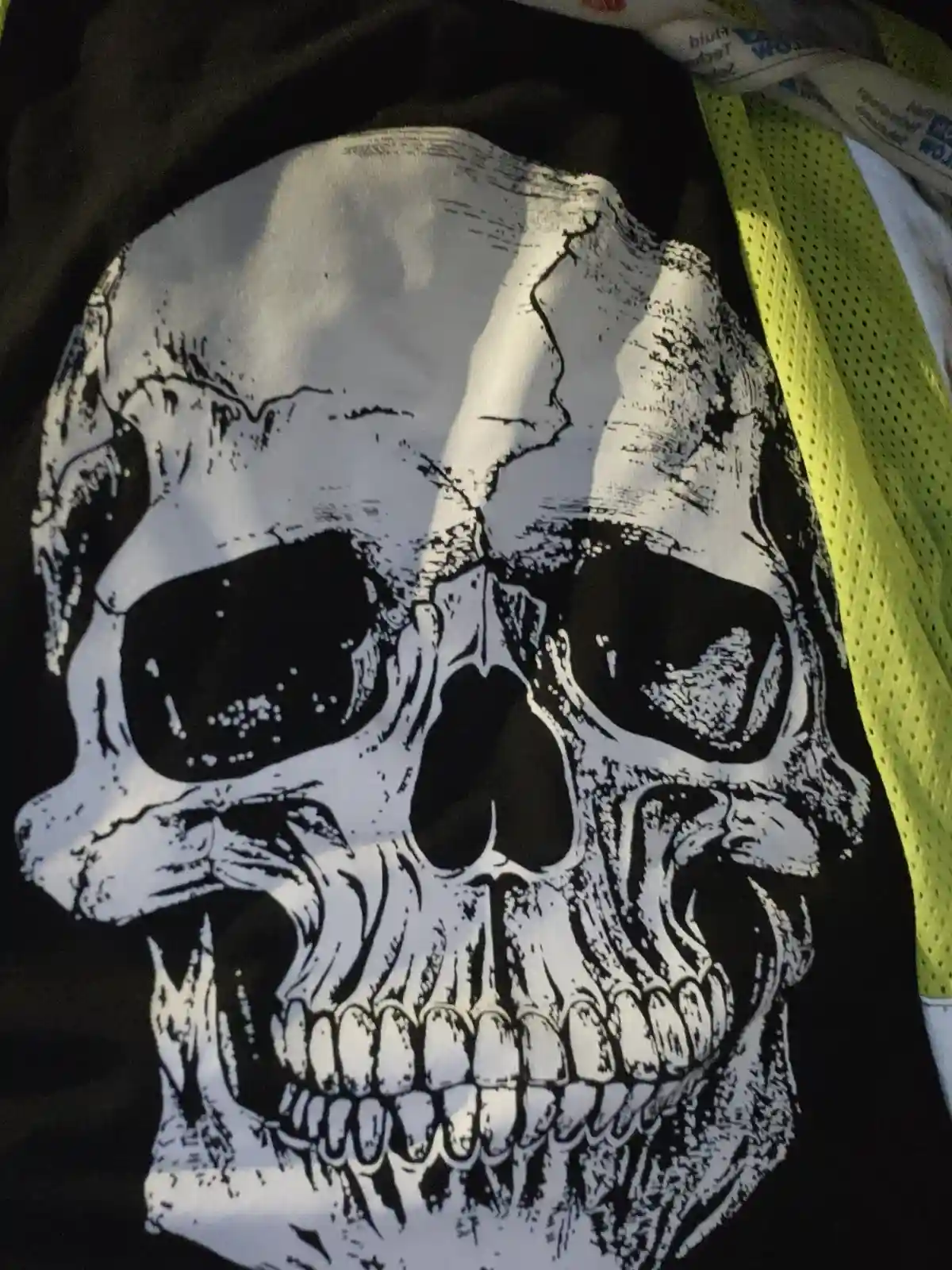 A close-up of a black t-shirt featuring a large, detailed white skull graphic with a cracked forehead. A bright yellow mesh…