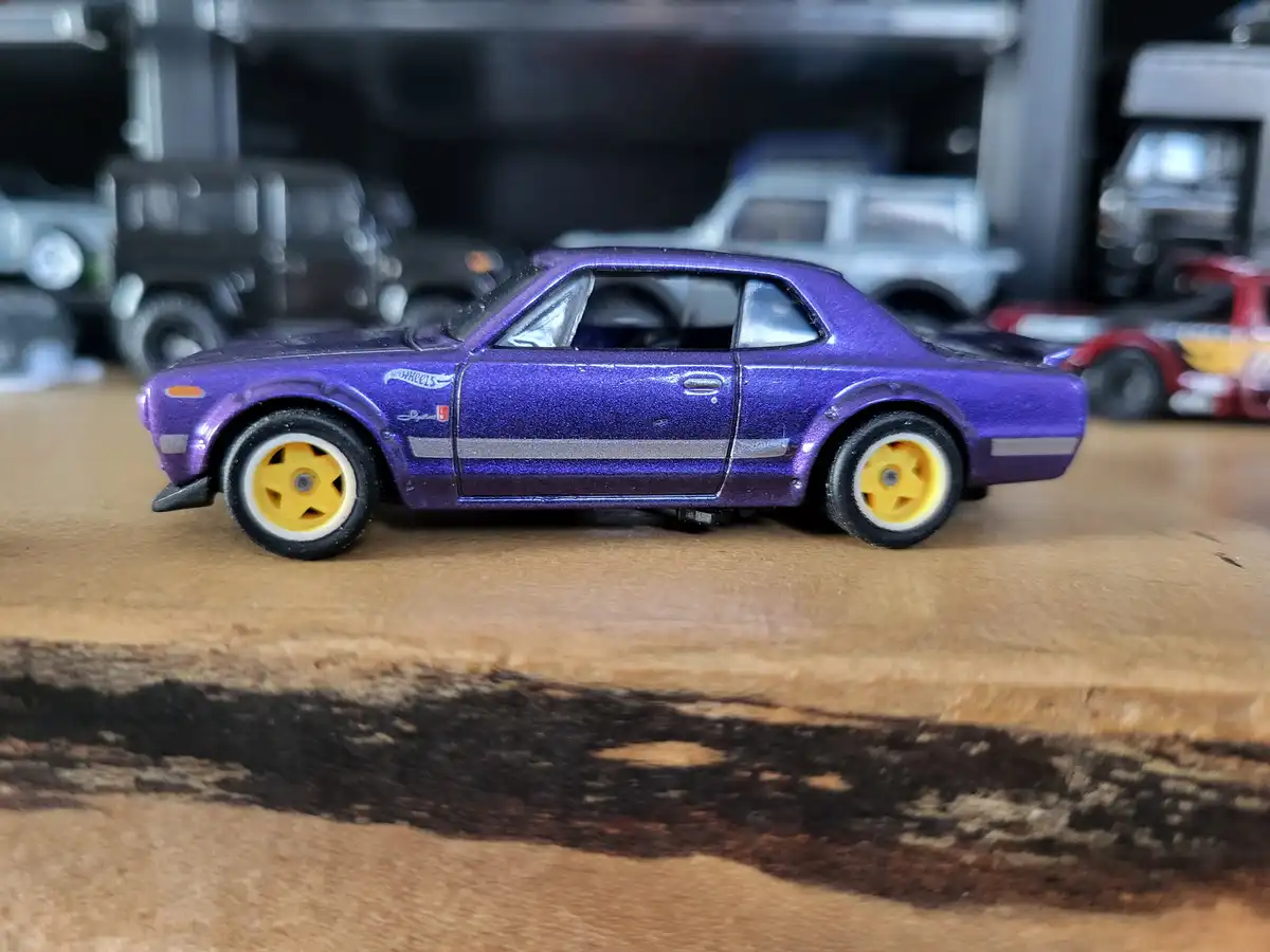 A purple glittery Hot Wheels toy car with yellow wheels sits on a wooden surface, with other toy cars blurred in the…