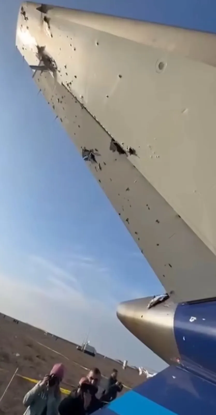 A low-angle shot shows the heavily damaged underside of an airplane wing, riddled with numerous bullet holes and large…