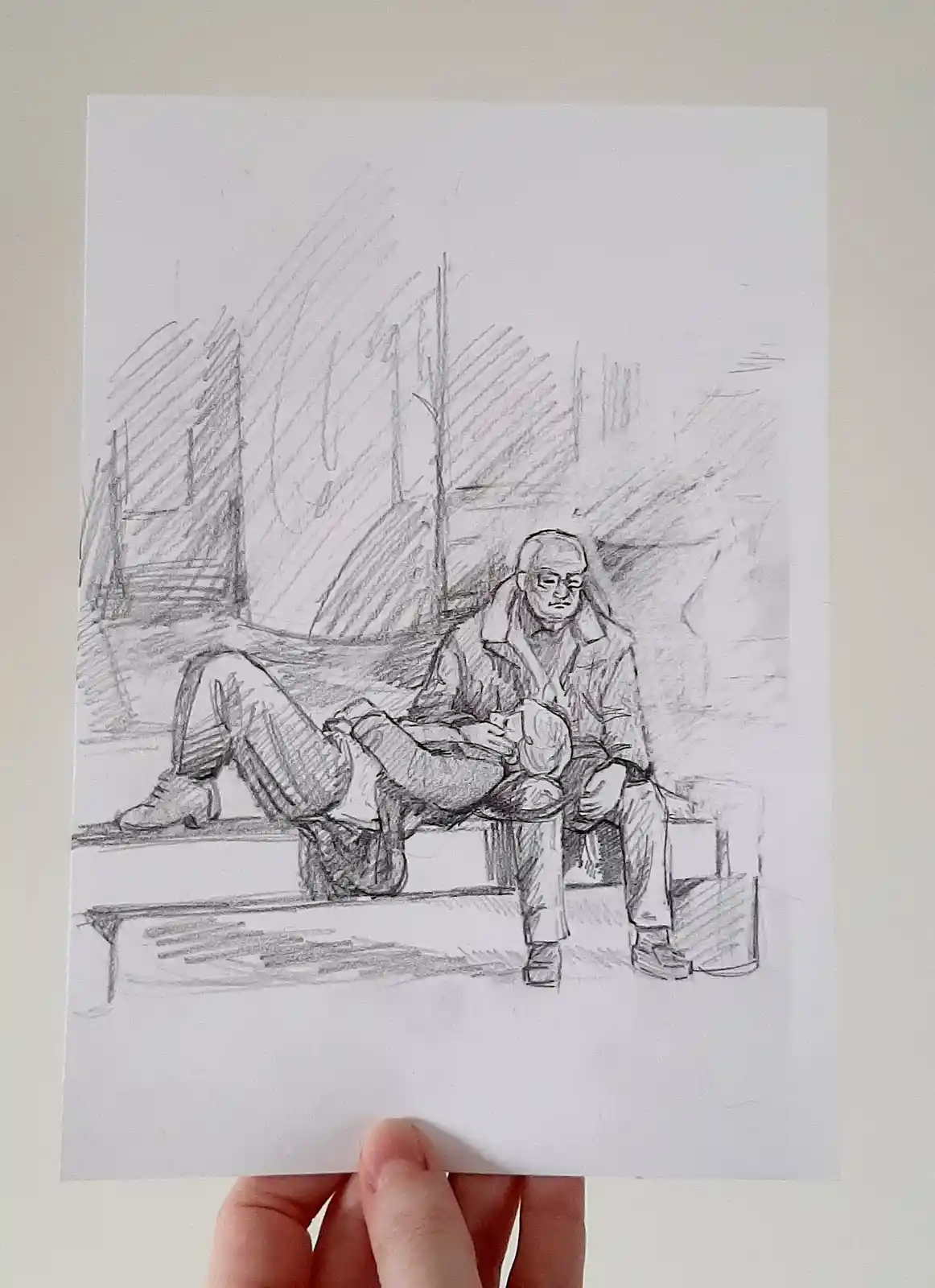 A hand holds up a pencil sketch depicting an elderly man sitting on a bench with his legs crossed, looking down at something…