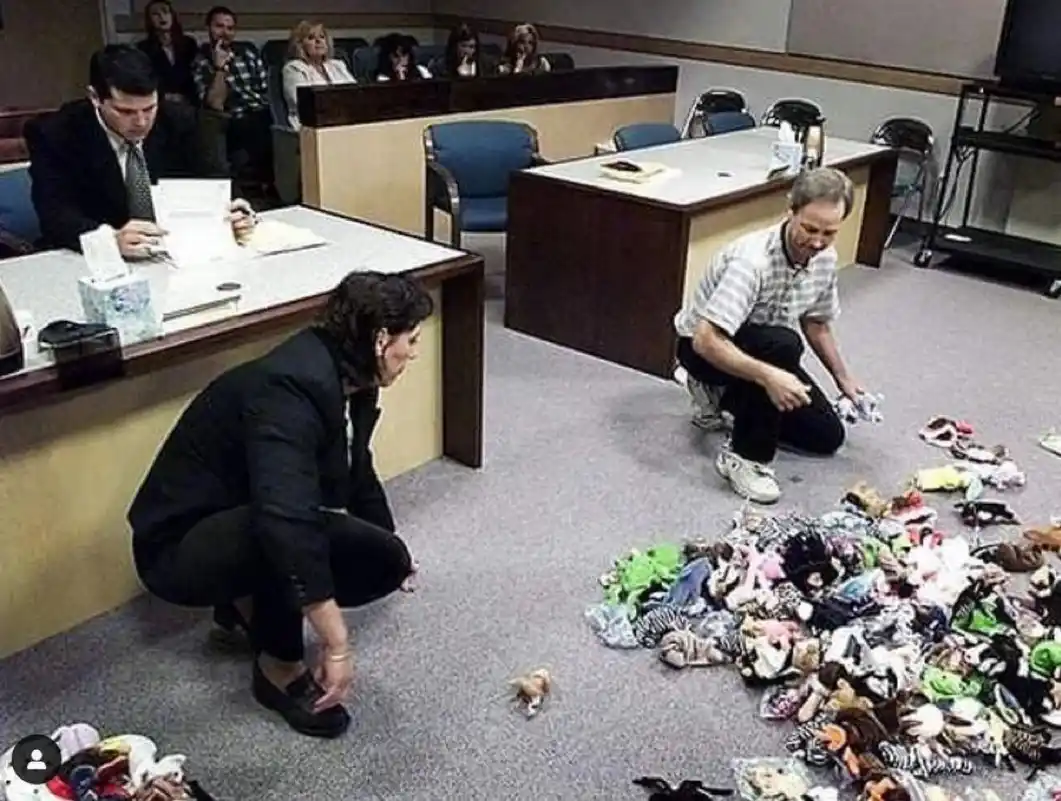 In a courtroom, two people kneel on the floor surrounded by a large pile of stuffed animals. One person, in a black suit…