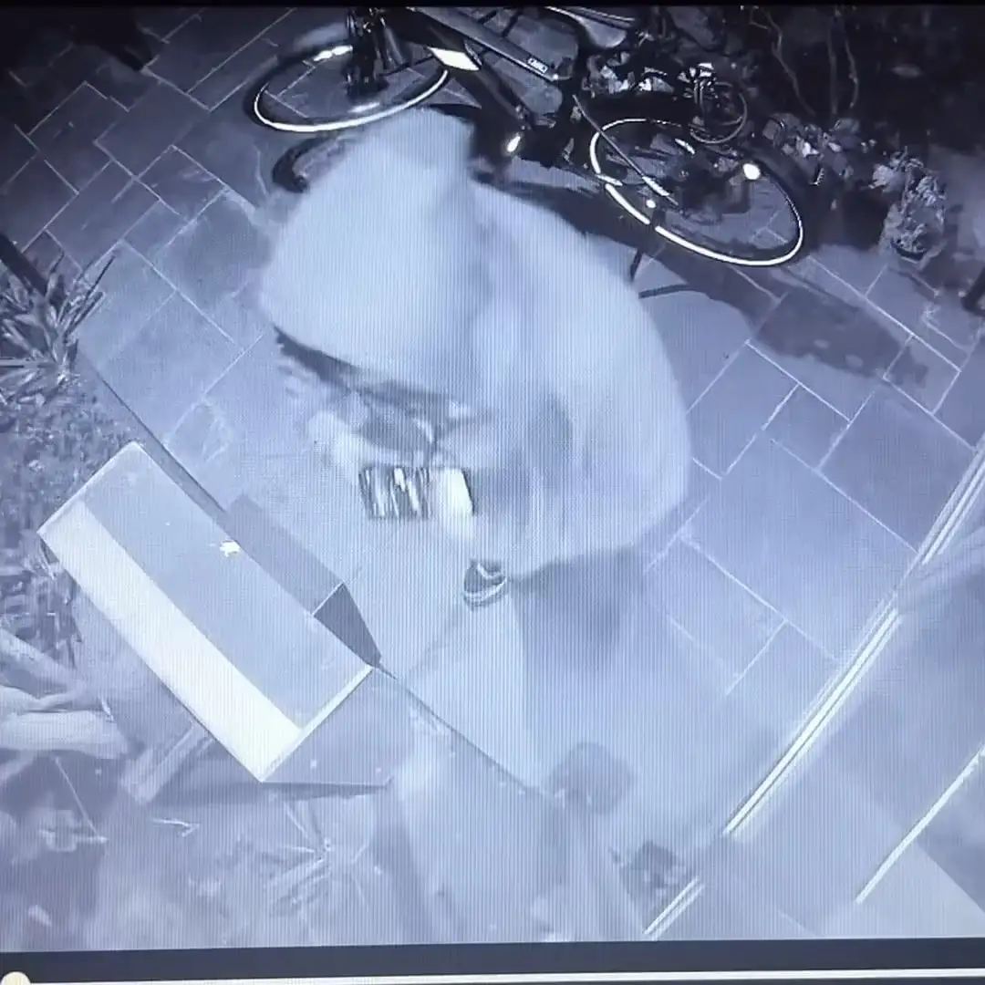 A blurry, low-resolution security camera image shows a person in motion, appearing to be bending or kneeling over a white…