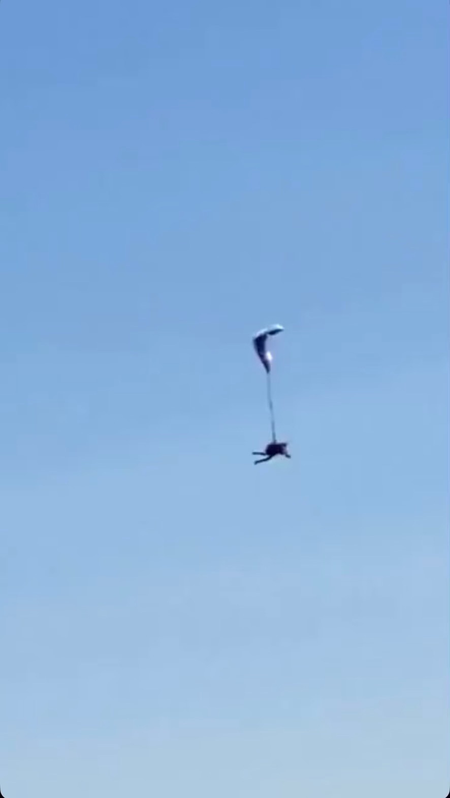 A person is suspended in mid-air against a clear blue sky, attached to a parachute that is open and catching wind. The…