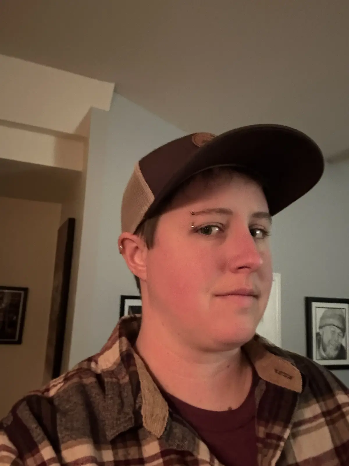 A person with short hair, wearing a brown and beige trucker cap and a plaid flannel shirt over a maroon t-shirt, looks…