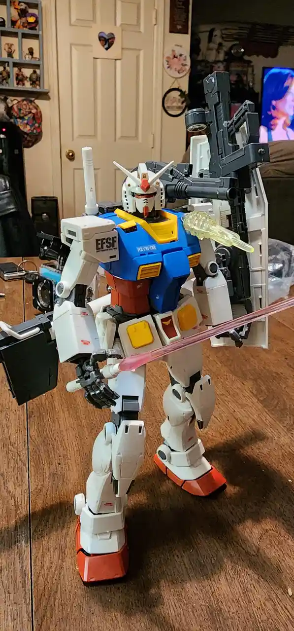A blue, white, and orange Gundam model, marked 'EFSF' and 'RX-78-02', stands on a wooden floor holding a pink beam saber and…