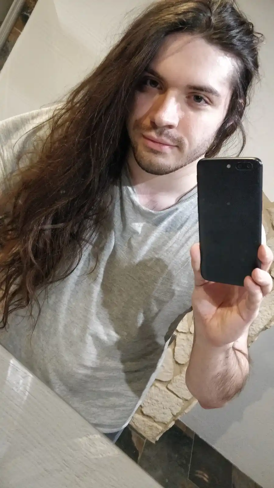 A man with long brown hair and a light beard takes a mirror selfie holding a black smartphone with its screen off. He wears…