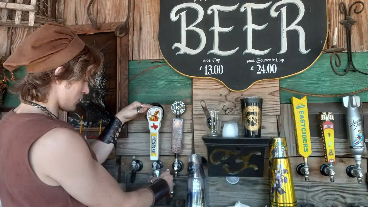 A person in a brown hat and sleeveless shirt pulls a beer tap at a rustic wooden bar with a large sign reading 'BEER' and…