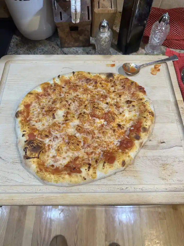 A homemade cheese pizza with a slightly charred crust sits on a wooden cutting board, with a spoon smeared with sauce…