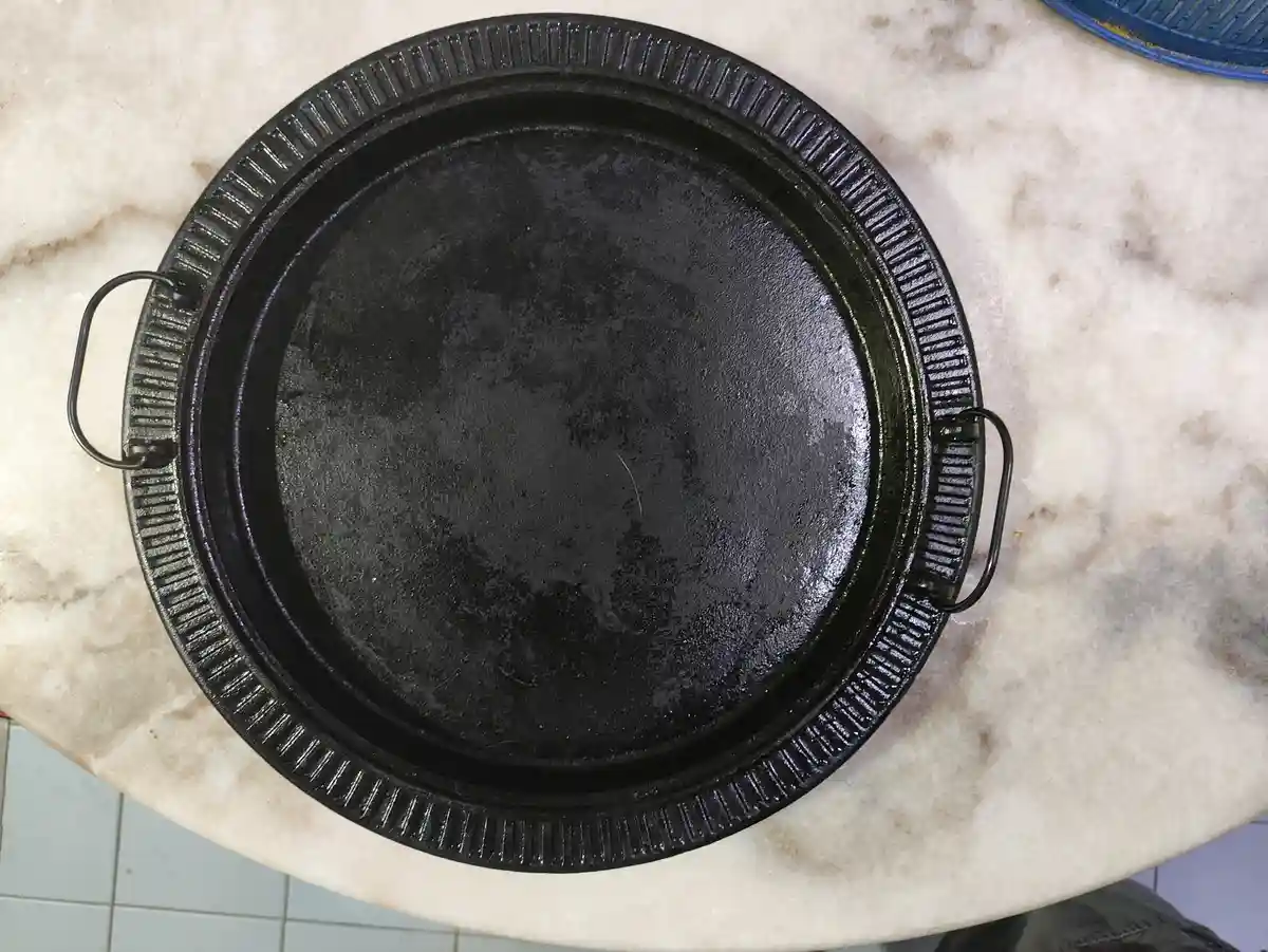 A top-down view of a black, round cast iron pan with two loop handles, resting on a light-colored marble countertop. The…