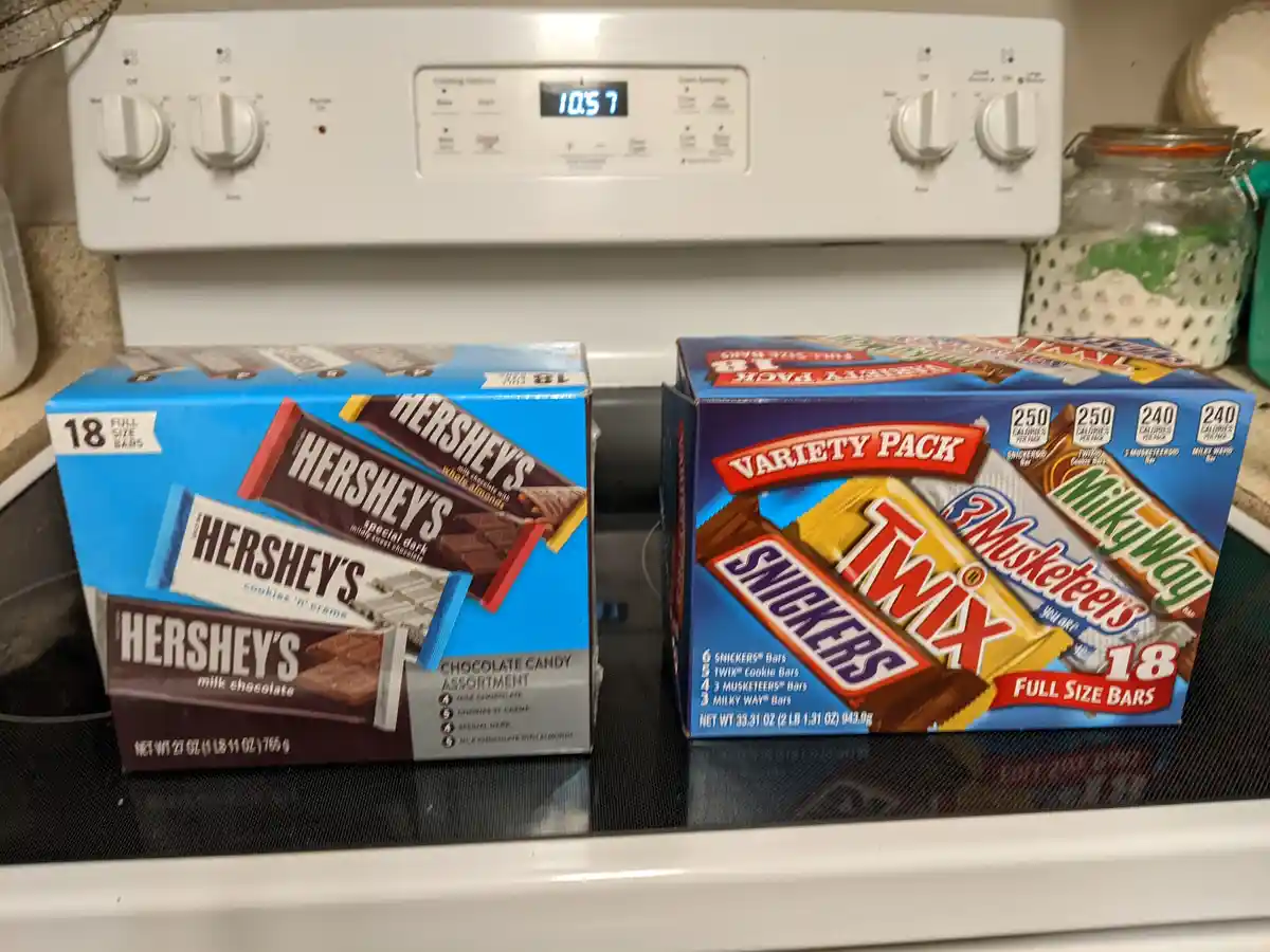 Two large boxes of chocolate candy sit side-by-side on a black electric stovetop, in front of a white oven with a digital…