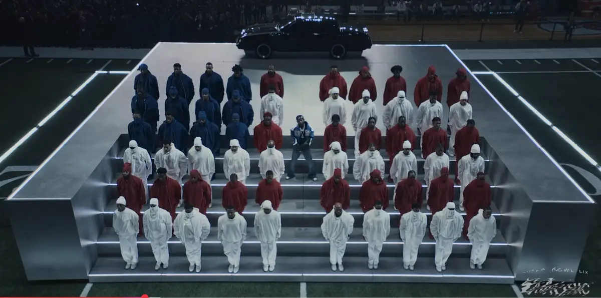 A wide shot shows a large group of men in red, white, and blue hooded outfits standing in rows on a multi-level metallic…