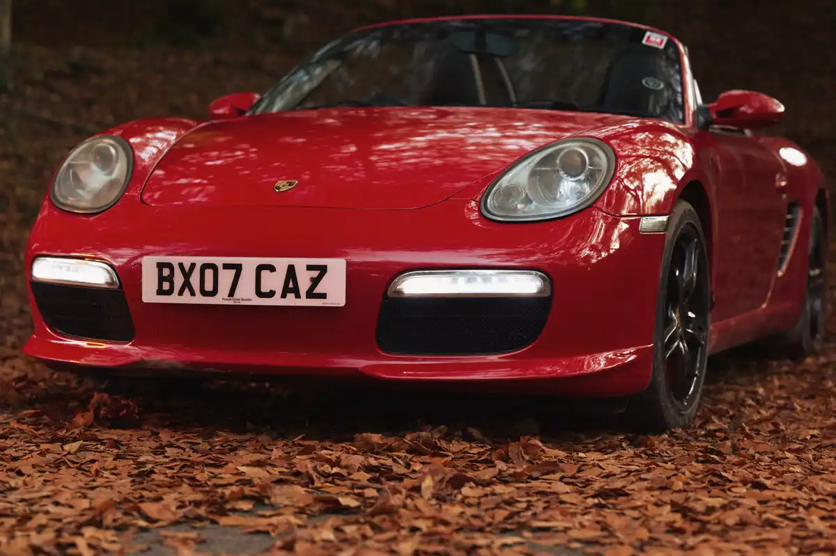 A red Porsche Boxster convertible is parked on a path covered in brown autumn leaves, with its front license plate clearly…