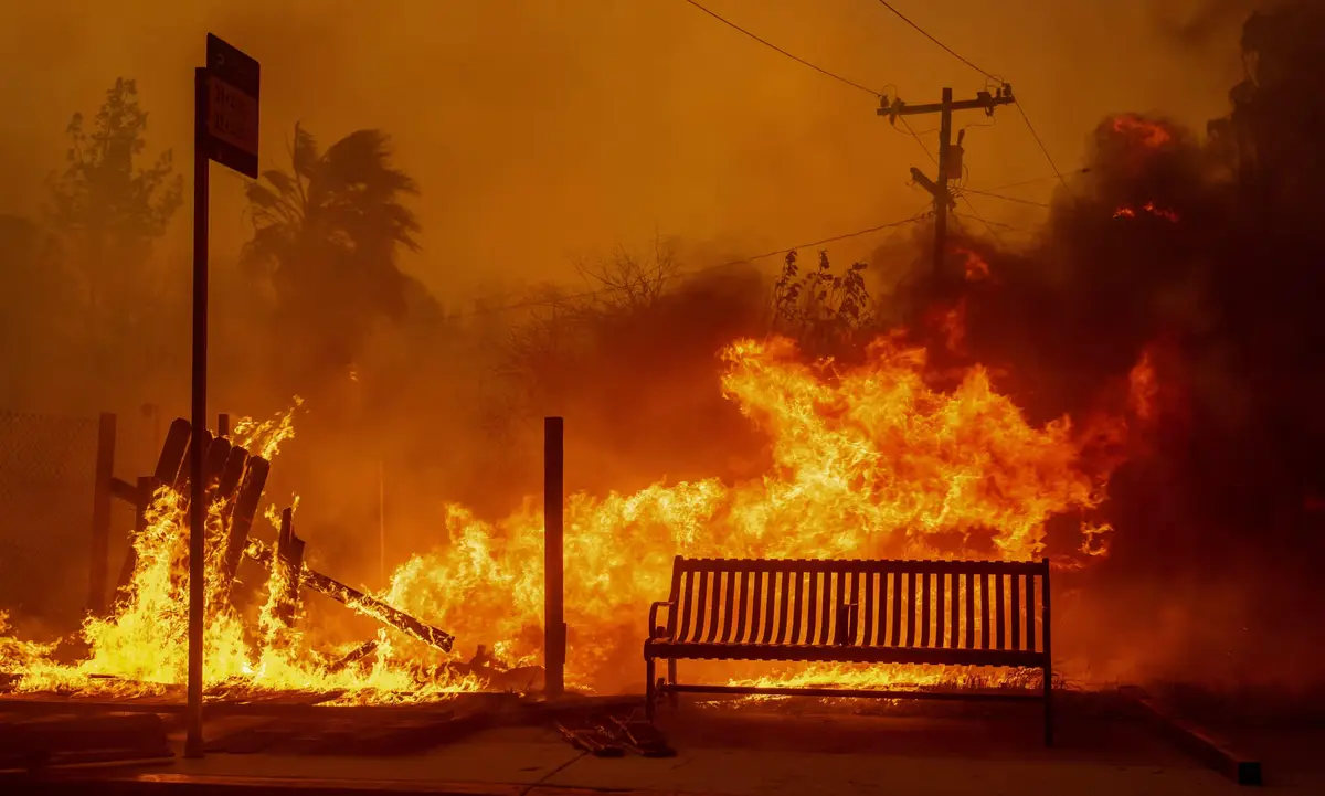 A public bench sits calmly in front of a raging wildfire, with flames and smoke filling the background. A bus stop sign is…