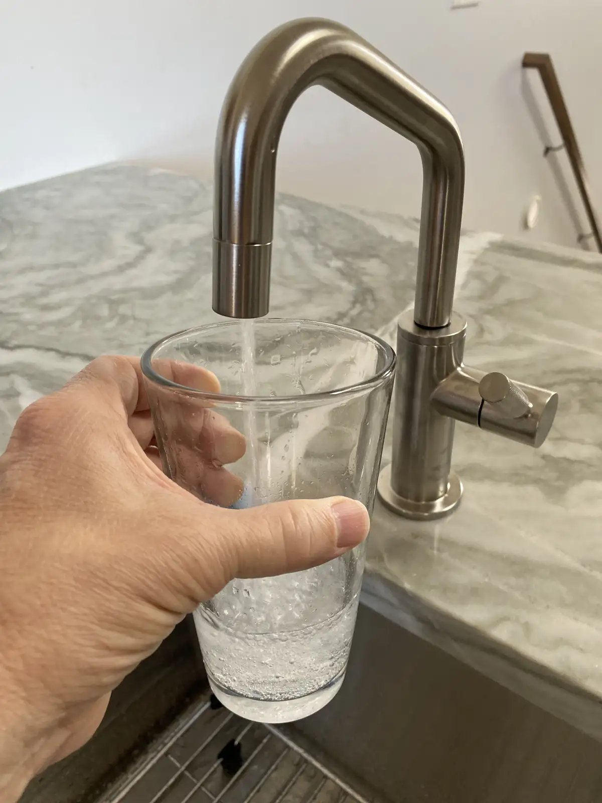 A hand holds a clear glass under a modern stainless steel faucet as water flows into it, set against a marble countertop…