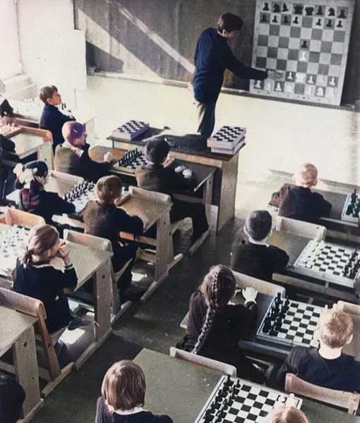 An overhead view of a classroom where children sit at desks with chessboards, while a teacher stands at the front pointing…